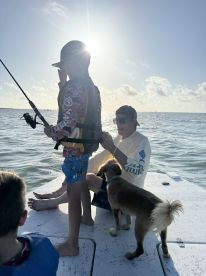 A young boy in a life jacket is casting a fishing line while enjoying a sunny day on the water, accompanied by an adult and a dog.