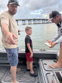 A young boy stands on a boat, focused on fishing, while an adult prepares to show him a catch. The scene captures a family fishing experience in a coastal environment.