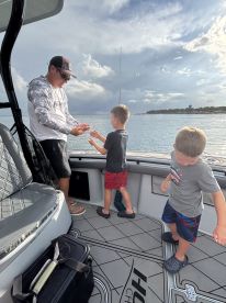 A young boy is excitedly learning to fish on a boat, while an adult demonstrates how to handle the catch. The scene captures a family-friendly fishing experience on calm waters.