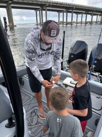 A fishing guide demonstrates bait preparation to two young anglers on a boat, with a bridge in the background, highlighting an engaging fishing experience.