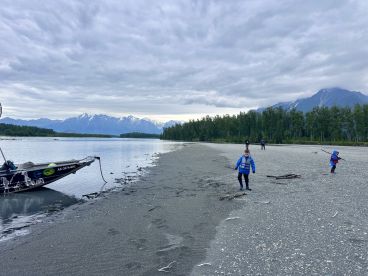 Two children explore the sandy shore near a fishing boat, surrounded by stunning mountain scenery in a tranquil river setting.