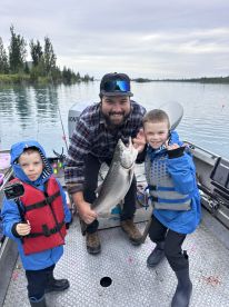 A family fishing trip showcasing a proud moment with a large trout caught on a scenic lake, featuring two young anglers and their father.