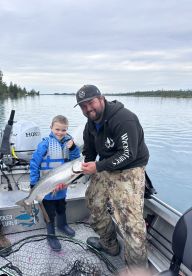 A young boy proudly displays a large trout alongside an adult angler on a fishing boat, surrounded by a serene lake environment.