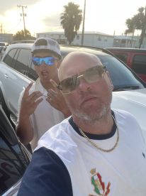 Two individuals posing for a selfie in a parking lot, with palm trees and vehicles in the background.