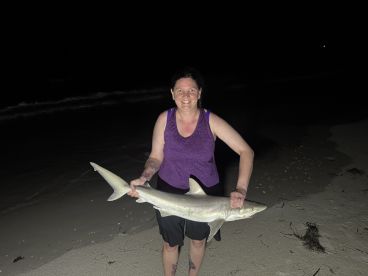 Angler proudly displaying a caught shark on the beach at night, showcasing a successful fishing experience.