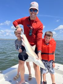 A proud father and his two children display a caught shark while enjoying a sunny day on the water, showcasing a family fishing adventure.