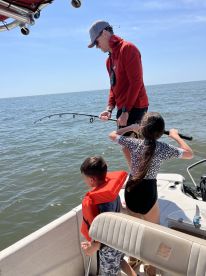 A family enjoying a fishing trip on a boat, with a child assisting an adult in reeling in a catch on a sunny day.