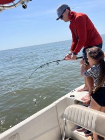 A young girl and an adult are engaged in fishing, with the adult helping her reel in a catch on a sunny day at sea.