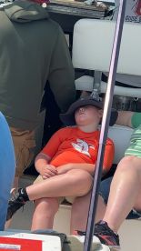 A young boy resting in a seated position on a fishing boat, wearing a shark-themed shirt and a wide-brimmed hat.