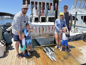 A family poses proudly in front of their catch, featuring a variety of fish including Snapper and Mackerel, displayed on a catch board at the dock.