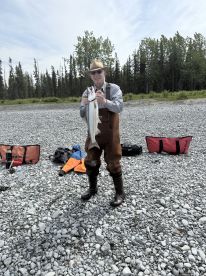 An angler proudly displaying a large fish on a rocky riverbank, surrounded by fishing gear and lush greenery in the background.
