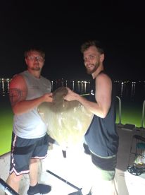 Two anglers proudly displaying a large stingray caught during a night fishing trip, illuminated by boat lights.