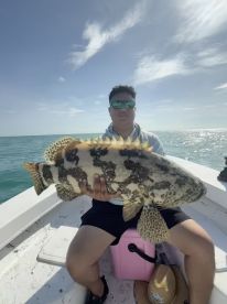 An angler proudly displaying a large Goliath Grouper while seated on a boat in clear waters, showcasing a successful fishing trip.