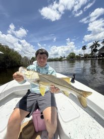 A young angler proudly displaying a Snook while fishing from a boat in a scenic waterway, surrounded by lush greenery and palm trees.