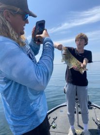 A young angler proudly displaying a caught Bass while another person captures the moment on a phone, enjoying a day of fishing on a calm lake.