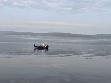 A fishing boat is seen gliding across a calm lake, surrounded by misty hills, creating a serene fishing environment.