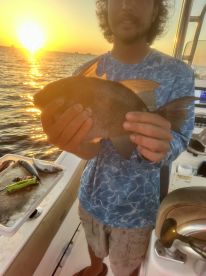 Angler holding a Snapper against a stunning sunset backdrop while fishing on a boat.
