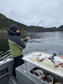 A young angler is actively reeling in a catch while fishing from a boat in a scenic coastal environment, with rocky cliffs and lush greenery in the background.