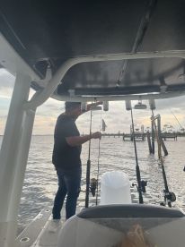 Angler actively fishing from a boat, with multiple fishing rods visible and a scenic waterfront backdrop.