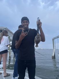 An angler proudly displaying a Flounder while fishing from a boat, with friends in the background enjoying the day on the water.