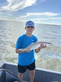 Young angler proudly holding a Speckled Trout while fishing on a boat in a coastal waterway.