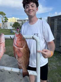 A young angler proudly displaying a large Grouper caught during a fishing trip, with a scenic backdrop of a coastal area.