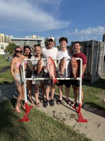 A group of six anglers proudly displaying their catch of Red Snapper on a catch board after a successful fishing trip.