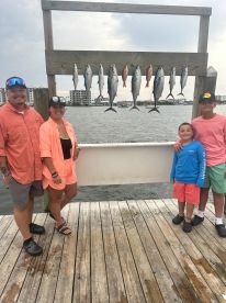 A family poses proudly in front of a catch board displaying a variety of fish, including Mackerel and Snapper, after a successful fishing trip.