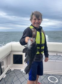 Young angler proudly holding a large Black Sea Bass while fishing on a boat, showcasing a successful catch.
