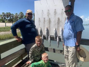 A family poses proudly in front of a catch board displaying several Speckled Trout after a successful fishing trip at the coast.