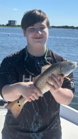 Young angler proudly holding a Redfish while on a fishing trip, with a scenic waterfront backdrop.