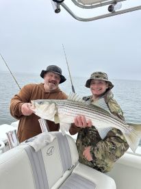 Two anglers proudly displaying a large Striped Bass aboard a boat during a fishing trip.