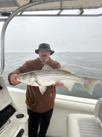 Angler proudly displaying a large Striped Bass aboard a fishing boat during a cloudy day on the water.