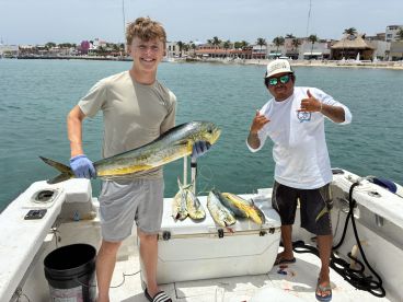 A young angler proudly displaying a large Mahi Mahi alongside a crew member on a fishing boat, with a vibrant coastal backdrop.