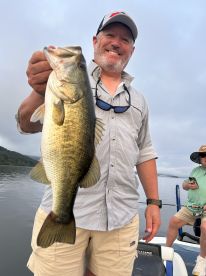 An angler proudly displaying a large Largemouth Bass while fishing on a calm lake, with another fisherman visible in the background.