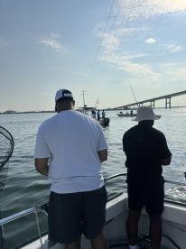 Two anglers are fishing from a boat, with a bridge in the background, showcasing a serene day on the water.