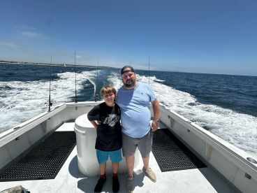 A father and son enjoying a day on a fishing boat, with fishing rods visible in the background and a scenic ocean view.