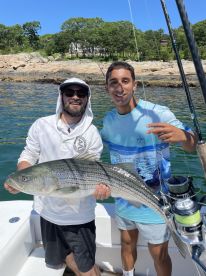 Two anglers proudly displaying a large Striped Bass while fishing on a sunny day in a coastal environment.