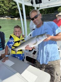A young boy in a life jacket proudly poses with a striped bass alongside an adult angler on a boat, showcasing a successful fishing trip.