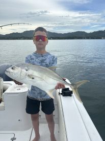 A young angler proudly displaying a large Jack Crevalle while fishing on a boat in a scenic coastal environment.