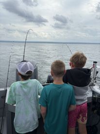 Three young anglers engaged in fishing on a boat, with rods bent as they reel in their catches on a cloudy day.