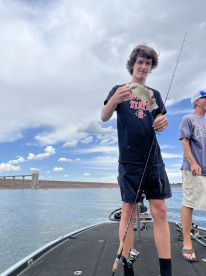 A young angler proudly displaying a caught fish while fishing on a boat, with a scenic backdrop of blue skies and water.