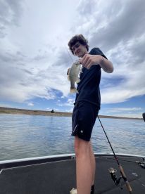A young angler proudly displaying a freshly caught bass while fishing on a calm lake, with a scenic backdrop of clouds and shoreline.
