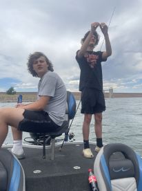 Two young anglers on a boat, one preparing to cast while the other relaxes, enjoying a day of fishing on a calm lake.