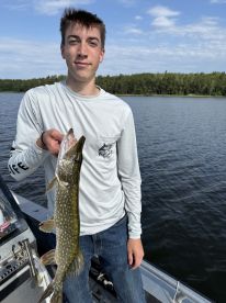 A young angler proudly displaying a Northern Pike while fishing on a serene lake, surrounded by lush greenery.