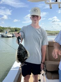 A young angler proudly displaying a Flounder while fishing on a boat, showcasing a successful catch in a scenic coastal environment.