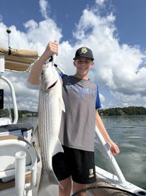 A young angler proudly displaying a large striped bass while fishing on a boat, surrounded by a scenic lake backdrop.