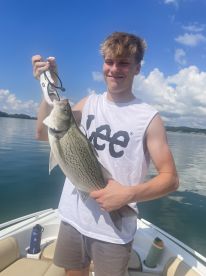 A young angler proudly displaying a striped bass while fishing on a calm lake, showcasing a sunny day and clear blue skies.