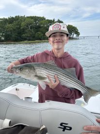 Young angler proudly holding a large Striped Bass while fishing on a boat, showcasing a successful day on the water.