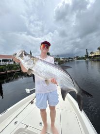 A young angler proudly displaying a large Tarpon while fishing from a boat in a scenic canal.